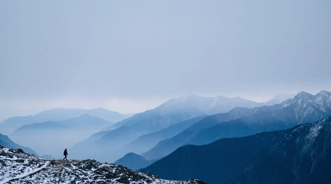 A lone walker on a high alpine ridgeline at first light, with layered mountain ridges receding into cold morning haze.